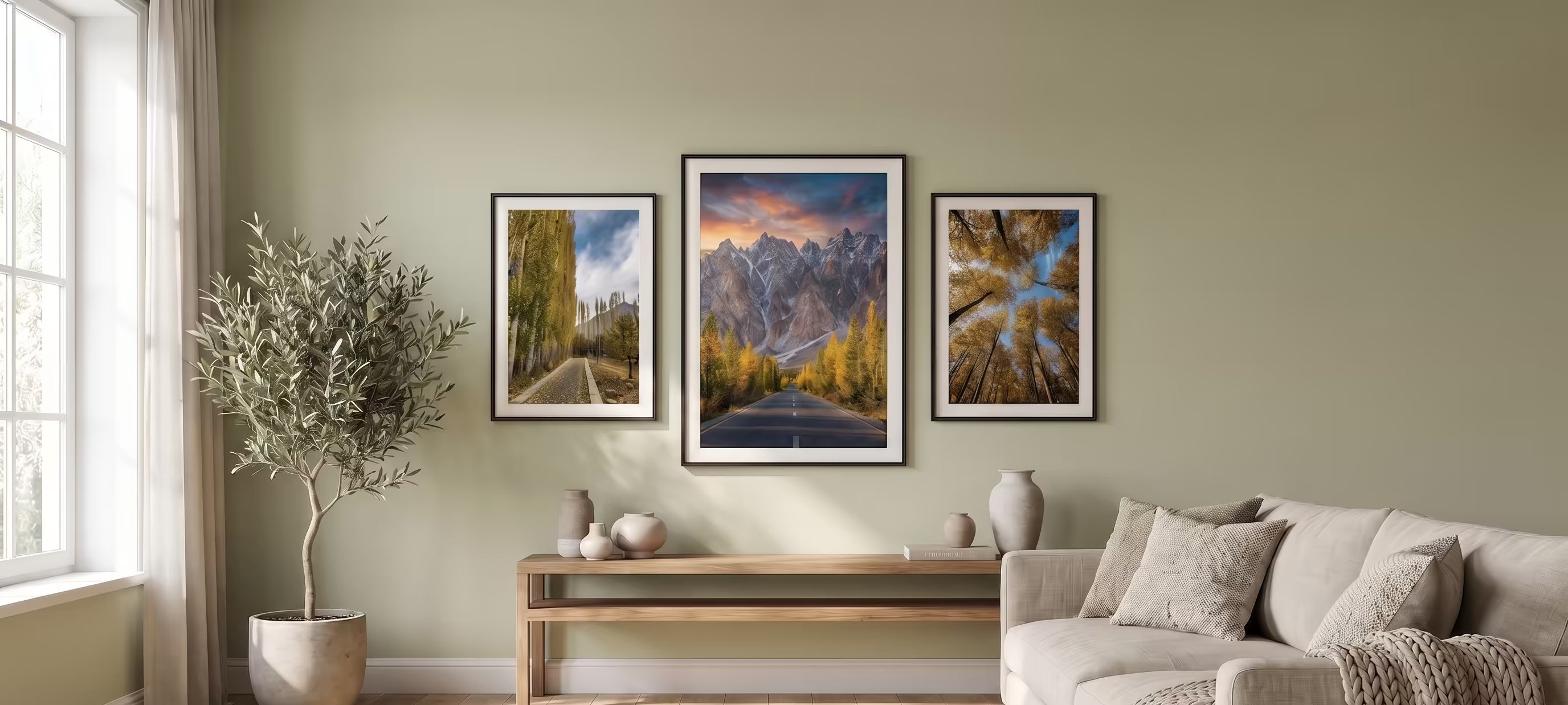 A styled living room scene showing three framed landscape art prints hanging on a sage green wall above a wooden console table and a light beige sofa. The prints depict a winding road through trees, a mountain range at sunset, and a forest path with colorful autumn foliage.